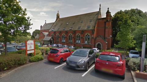 A large red brick building with a sloped roof and arched windows and a car park in front with a number of cars in it, surrounded by bushes
