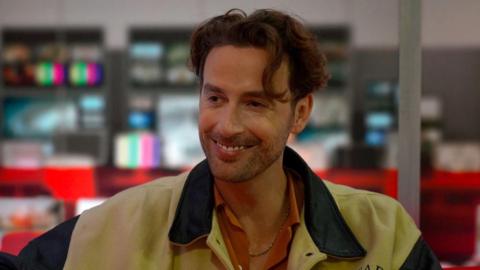 A photo of an actor called Tim Renouf. He is in a ˿ studio. He is looking off towards the right and smiling. He has brown curly hair. He is wearing a black and cream jacket and an orange shirt.