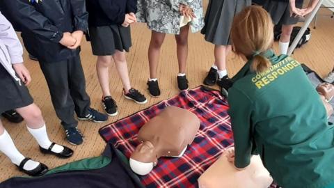 A woman attempting CPR on a simulation dummy. There are people watching the demonstration, and the lower halves of school children can be seen in the image. Their faces are not visible. There is another dummy also lying on the floor and a plaid blanket also on the floor.