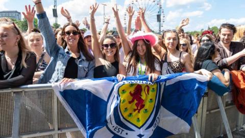 A group of young girls in festival wear standing at the barrier holding a saltire flag with Scotland emblem on it.