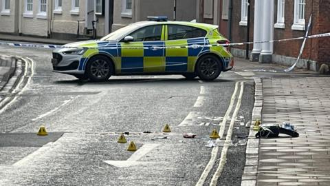 Police cordon and car at a crime scene in a residential street. Several yellow markers have been placed on the road next to debris. 