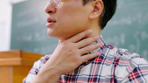 A man holds his throat as he struggles to speak to a room of people attending a lecture he is giving