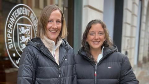 Jen and Sarah are in dark jackets stand on a wet pavement next to a large window with the words “Superintendent” and a crest logo. A stroller with a child seated inside is positioned in front of them. The street and other buildings are visible in the background.