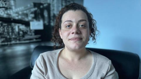 Kimberley pictured in her new home. She is sitting on a black leather chair, and a blue wall can be seen in the background. She has her hair tied up and is wearing a grey jumper. 
