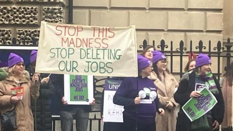 Protesters standing in a line wearing purple Unison hats. They are holding up signs and placards, the biggest of which reads: 'Stop the madness, stop deleting our jobs'. They are standing in front of black railings and a building.