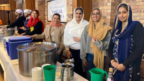 Five women stand in a line behind a table which has jugs, pans, boxes and bowls on top of it. All the women have their heads covered and are smiling. Two men can be seen in the background.