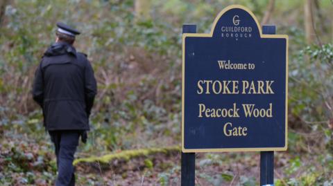 A police officers walks away from a sign into a wooded area. The sign reads "Welcome to Stoke Park Peacock Wood Gate".