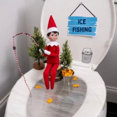 Elf sitting on toilet seat holding a fishing rod which is dangling into the loo. The toilet is covered in cling wrap and has little fir trees on it and a sign saying "ice fishing". 