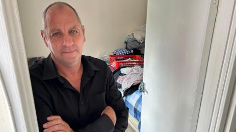 Colin Hanwell is standing in a bedroom with items on a bed behind him. He is leaning on a door frame with his arms crossed. He is wearing a black shirt and has short spiky grey hair around the sides an is bald on top. 