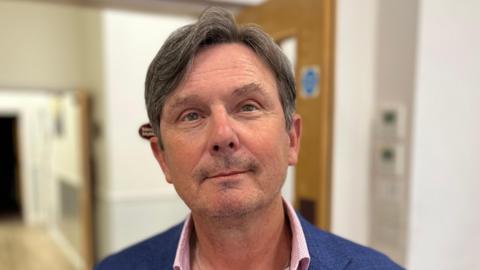 A man called Andy Sloan. He has neatly trimmed dark-greying hair. He is stood in front of a double doorway and is wearing a navy blue jacket and a pink shirt.