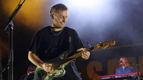 Sam Fender performs onstage for day two of the 2025 Pilgrimage Music & Cultural Festival at The Park at Harlinsdale Farm. He is playing a green electric guitar and is smiling down at his instrument. He is wearing a black T-shirt and silver chain. A keyboard player is behind him on stage.