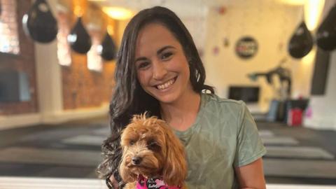 Leanne Lucas, who has long, dark brown hair, smiles while holding a therapy dog in a yoga studio.