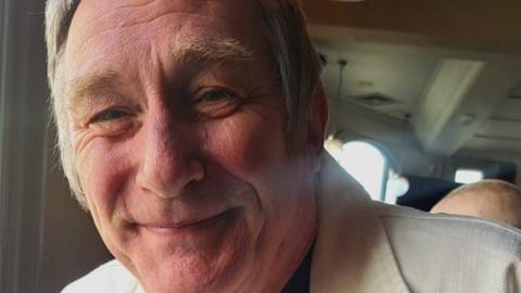 A close-up of a man in his 50s. He has a slightly red face and is smiling with his mouth closed. He has sideburns and a white collared shirt-jacket.
