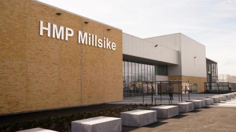 The outside of the jail. Light brown brickwork to the left with the sign HMP Millsike in large white letters, while to the right is a glass-front entrance.