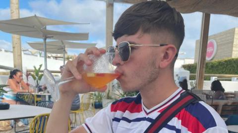 Joel Sheldon is holding a drink to his lips as he sits in an outdoor seating area. He is wearing sunglasses and has short brown hair. He is wearing a red, white and blue T-shirt.