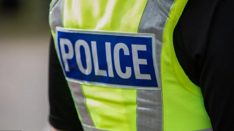 The back view of a police officer wearing a yellow vest with the words police in bold white lettering against a blue rectangular background.