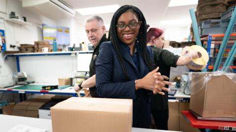 Kemi Badenoch in a factory in Brentwood with two employees behind her as they box up goods.