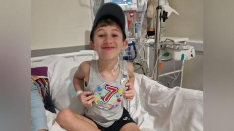 A boy sits on a hospital bed. He is wearing a grey vest, black shorts, a black baseball cap and a large multicoloured badge that says "7 Today" on it. He has brown hair and he is smiling.