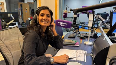 A woman wearing a black shirt and headphones while smiling towards the camera. She is sat next to a purple microphone with white writing on it reading "BBC Radio Kent".