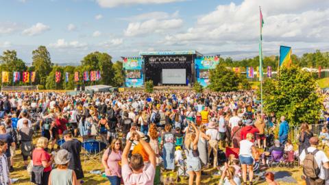 A large crowd begins to gather in front of the main stage in a field in the sunshine. 