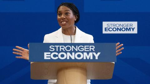 Conservative Party leader Kemi Badenoch gives a speech from behind a podium reading "stronger economy" in front of a blue backdrop.
