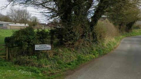 A view of a road lined by a hedge with a sign saying Upton Scudamore on the left verge