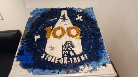 A Lego model of Stoke-on-Trent's centenary logo, which is made up of hundreds of grey, black, blue and white plastic bricks. Models of a Spitfire plane and a bottle oven can be seen in the middle, alongside the number 100 in gold Lego pieces and the word "Stoke-on-Trent" written in white along the bottom.