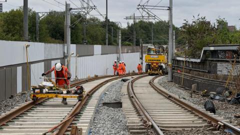 Five construction workers are on a tram track wearing orange high-vis jackets, one of them is pushing a yellow piece of machinery, the others are standing in the distance next to a vehicle.