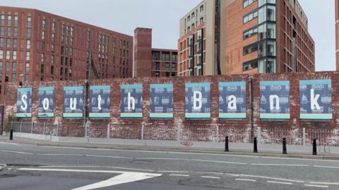 A brick wall with the words 'South Bank' spelt out on blue boarded signs on it. Brick buildings are behind it.