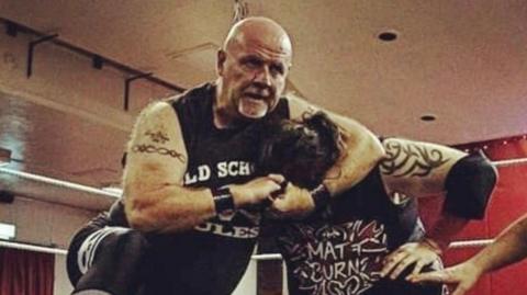A man wearing a black wrestling suit is holding another man in a headlock. Both men have tattoos on their arms. They are indoors, in a wrestling ring with the ropes visible in the background.