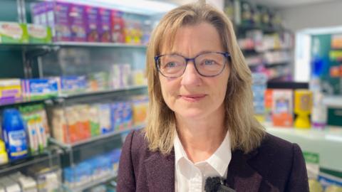 Pharmacist Georgina Frith wearing a purple jacket and white blouse standing in front of shelves stacked with medicines