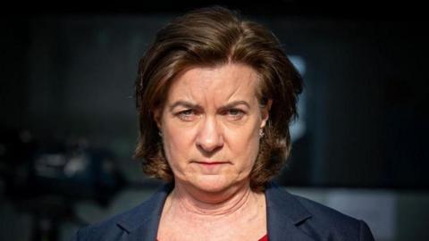 A head and shoulders framed picture of Eluned Morgan in artificial light, with a blurred and very dark background behind her. She has a very serious expression on her face.