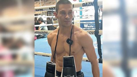 A man standing in front of a boxing ring, which has a blue floor. He is topless with a pair of black and white boxing gloves hanging around his neck. The image cuts off at the waistband of his black pair of shorts.