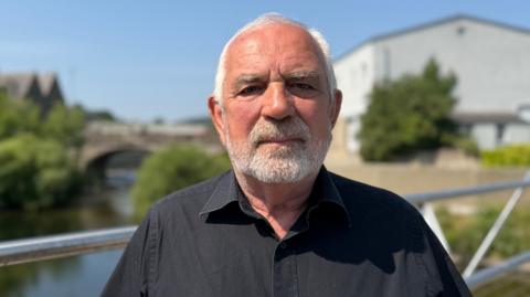 A man with grey hair and a grey beard in a black shirt stands on a bridge with a river, trees and a white building behind him. The background is blurred but there looks like another bridge over the water behind him.