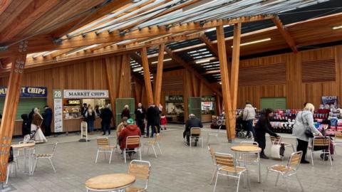 Great Yarmouth's market hall, a wooden structure with zinc roof. In the foreground is seating and tables, with people milling about inside the market building. Stalls can be seen in the backdrop of the building.