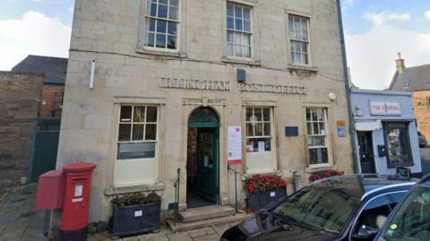 A view of a cream-coloured brick building which serves as Uppingham Post Office. A red post box is visible out front.