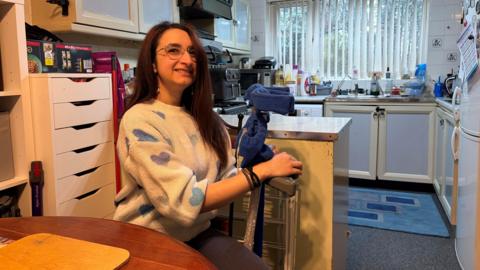A woman sat down inside her home holding a pair of crutches. She is looking at the camera and smiling. She has long dark hair and is wearing glasses and a white sweater with blue and purple hearts on it. Her kitchen is behind her.