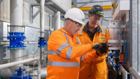Two men in orange body suits inside a water treatment works site.