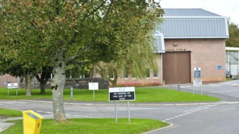 A picture of the outside of the prison in devon. There is a grass verge and signs surrounding the building. There is a large tree to the left of the image. 