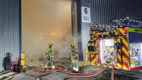 Two fire fighters and a fire engine outside a industrial unit, which has a sign that says "6 Bulky Waste" on it