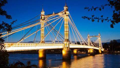 Albert Bridge is lit up at dusk with light bulbs across the spans of the structure and a turquoise sky behind. Lights are reflected onto the River Thames below.