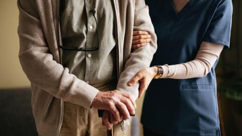 An elderly man holding a walking stick. A nurse wearing blue scrubs is holding his arm. 