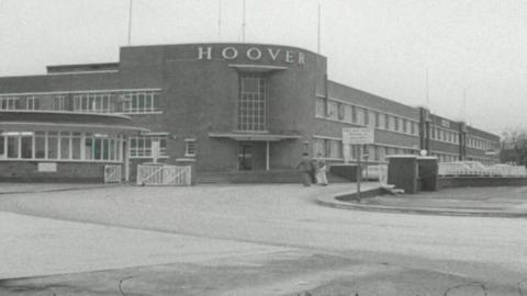 A general view of the Hoover factory. The brown building has 'Major Appliance Division' on two sides of it in white lettering. The entrance building is rounded with steps leading up to it. Two cars can be seen in the courtyard in front of it, as two people walk past on the street in front of it. 