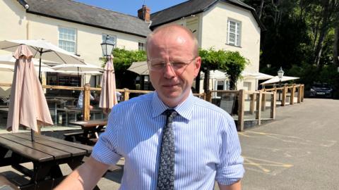 Matthew Cooke pictured outside the Malvern Hills Hotel. The two-storey building has been painted cream with a grey slate roof. He is wearing a pinstripe blue shirt with a grey tie. He has very short blond hair and thin wire glasses.
