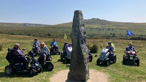 Eight people in mobility scooters in a semicircle around a large triangular rock. The grass is green. There is a hill in the background. The sky is a light blue. 