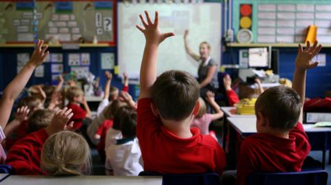 Children sit in a primary school classroom with several pupils raising their hands while a teacher stands at the front near an interactive whiteboard. School displays and learning materials can be seen around the room.