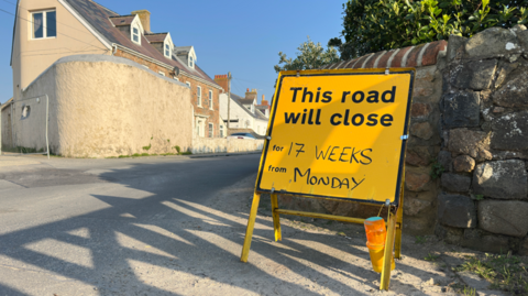A picture of a road sign. It is a roadwork sign yellow in colour. It says "This road will close for 17 weeks from Monday. There is a house pictured in the distance