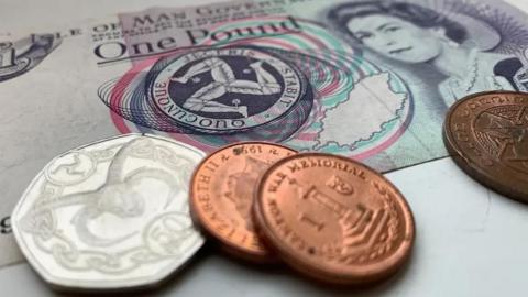 Manx cash and coins, a blue and pink £1 note on a table with a sliver 50p sitting next to it alongside three copper 2p pieces.