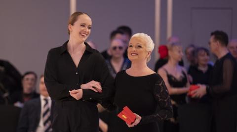 A woman, right, wearing a black dress and holding three red medals.
