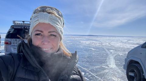 Anya smiles at the camera in a selfie. She is wearing a thick black coat and scarf, and a white knitted headband. There are a couple of 4x4 vehicles behind her, on a huge expanse of frozen lake. It is very sunny, with blue skies. 